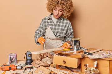 Fototapeta na wymiar Horizontal shot of professional female woodworker concentrated down paints wooden plank in workroom repairs old furniture wears checkered shirt overalls and protective gloves involved in carpentry