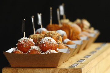 Deep fried farmers cottage cheese dumplings with various sprinkles, a sweet dessert served at a street food festival. Dark background, no people.