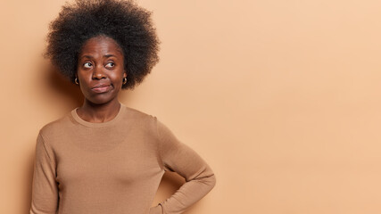 Fototapeta na wymiar Horizontal shot of thoughtful Afro woman with curly bushy hair focused tries to find solution of problem purses lips dressed in casual jumper isolated over brown background copy space for your text