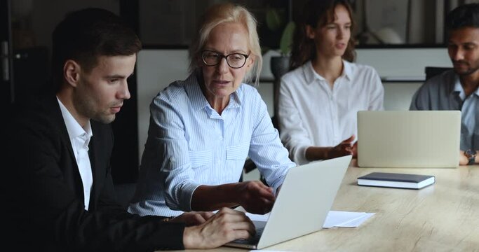 Mature female colleague helps to new employee with computer corporate software app in modern office, brainstorming ideas, explain online business services, share work moments, discuss online project