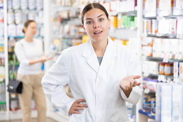 Young female pharmacist in uniform posing against backdrop of shelves with products in pharmacy