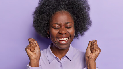 Charming optimistic African woman feels joyful smiles toothily shakesclenched fists keeps eyes closed and triumphs over something got success on work isolated on purple background. Yeah finally I won