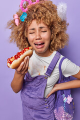 Upset crying curly haired pregnant woman eats delicious hot dog has changeable mood dressed in white t shirt and overalls carrie net bag isolated on purple background. Pregnancy and nutrition concept