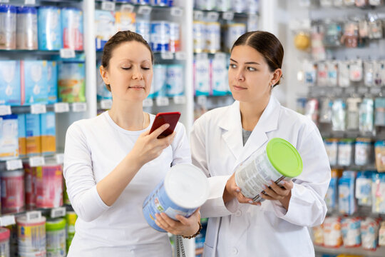 Woman pharmacist in medical uniform helps woman shopper in casual clothes find baby formula from photo on internet