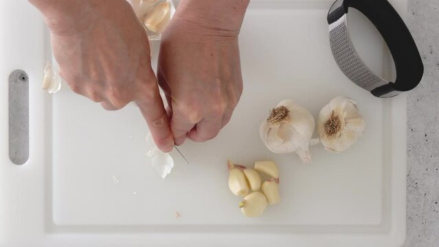 A woman presses fresh garlic using a garlic press tool on white cutting board on the kitchen table