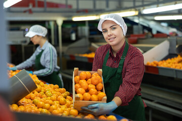 Happy young female worker of citrus sorting factory showing ripe selected mandarin oranges packed in wooden box © JackF