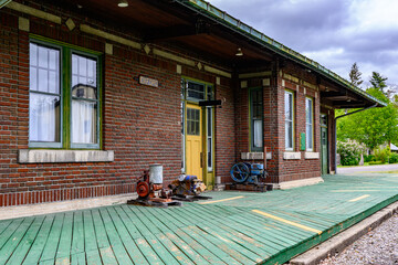 An old railroad station in now a museum in Oxford in Upstate NY.  Old antique transportation and train equipment sit outside for people to see.  Green wooden decking around brick building.
