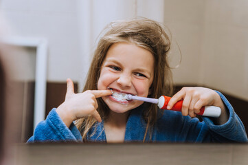 A little girl teenager, a child in a bathrobe cleans her teeth with a toothbrush while looking at herself in the mirror with reflection in the morning. Photography, close-up portrait.