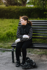 Fototapeta premium A schoolgirl girl, a child athlete with a plaster on her arm after a bone fracture sits on a bench in the park with roller skates, missing the ride. Photography, portrait, sickness, sport concept.