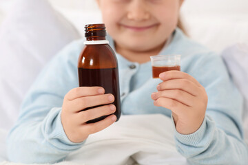 Closeup view of girl holding bottle and measuring cup with cough syrup in bed