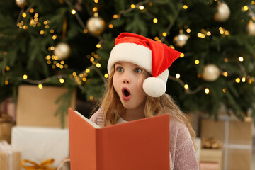 Emotional little girl in Santa hat with book near tree and gifts at home. Christmas atmosphere