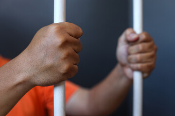 Close up of hands of prisoner in orange shirt inside the bars of a prison 