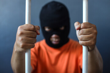 a prisoner in orange shirt and black mask inside the bars of a prison 