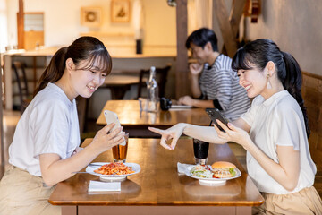 カフェでスマホを見ながら話す・おしゃべりする笑顔の若いアジア人女性たち
