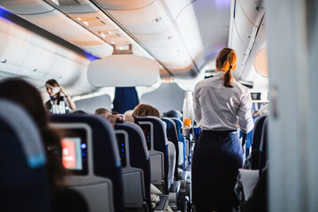 Interior of airplane with passengers on seats and stewardess in uniform walking the aisle, serving people. Commercial economy flight service concept - 604745576