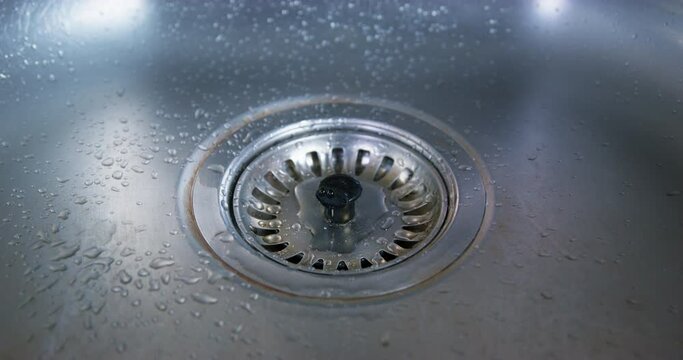 Close-up water from faulty kitchen faucet dripping into sink with plug drain. Outdated plumbing broken faucet leaking. Shortage shortage of drinking water saving utility bills. Repair of faulty faucet