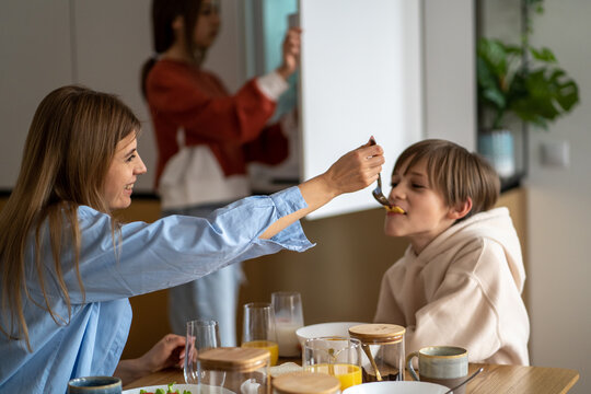 Happy mother feeding child son, having fun while enjoying breakfast together before school, sitting at kitchen table. Family mom and kids bonding over mealtime, mum eating with children in morning