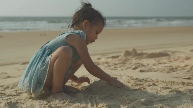 The girl sits on the sandy shore and builds a sand castle - Happy little girl plays with sand on the beach in the background at the blue sea at sunset.