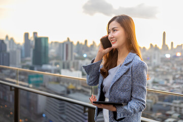 Fototapeta premium Young Asian business woman in formal suit is phone calling customer while standing outside the skyscraper building for marketing, connection corporate work, real estate, housing and urban development