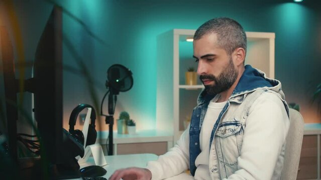 Happy young man in denim vest and white sweater works on a creative project on his computer in a vibrant home office studio. He is surrounded by colorful LED lights, black gadgets, and green plants.