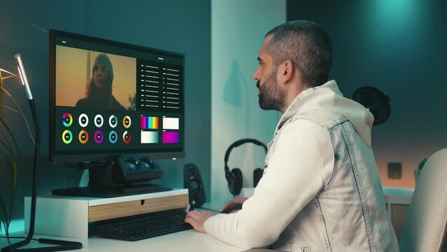 Young blogger edits a photo or video on his computer using color grading tools. He works in his cozy home office with black equipment and teal and orange lights. He looks creative and inspired.