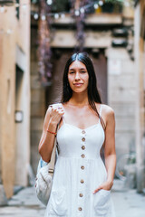 Hispanic woman with backpack holding hand in pocket of white dress and looking at camera while standing on blurred background of street of Barcelona, Spain