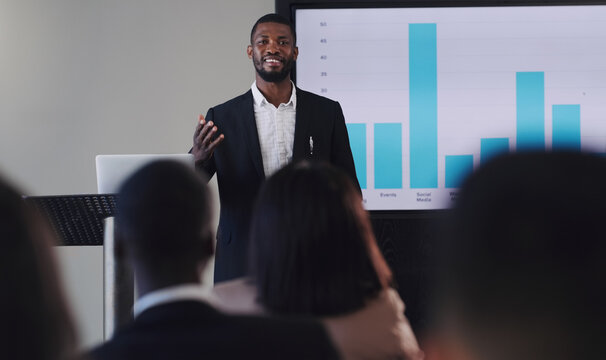 Black Man, Presentation And Speaker With Business Graphs At Seminar, Workshop Or Training. Men And Women Crowd At A Conference For Learning, Knowledge And Financial Growth Or Development Discussion