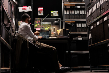 Detective studying crime scene surveillance photos on laptop screen, sitting at desk near evidence...