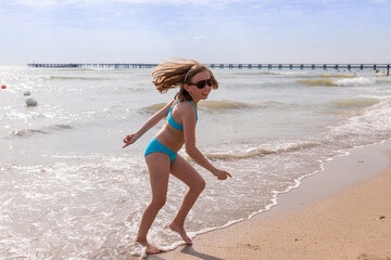 Fototapeta premium girl has a swimsuit, having fun alone on the sand in summer by the sea. female child dancing by the water while on holiday or vacation.happy and smiling girl performs gymnastic, dance exercises