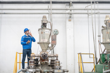 white american engineer standing in charge and use radio communication holding a list of items in the industrial plant There are machines working to produce steel and plastic works.