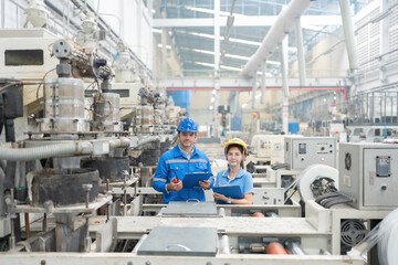 American engineers and Asian men and women, smart looking, standing with confidence, holding list notes, in plastic industry factory. There is a working machine Wear uniforms and helmets.