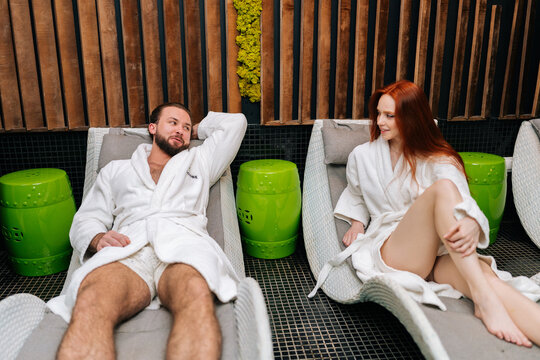 Portrait of cheerful young couple in white bathrobes talking lying on comfy loungers by poolside beauty salon. Handsome man and redhead woman enjoying relaxing after spa massage treatment.
