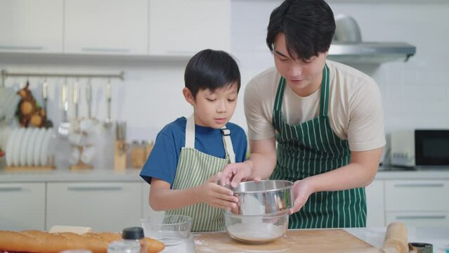 Happy asian father teaching little son sifts flour through sieve in glass bowl for cooking or baking enjoying weekend in kitchen room at home together. Family time concept