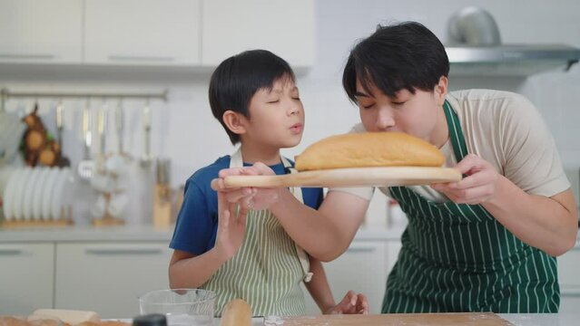 Happy asian father and son smelling of freshly baked loaf of bread on wooden tray in kitchen room at home. Bread homemade. Family time concept