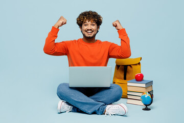 Full body young teen Indian boy student wear casual clothes sit near backpack bag pile of books use laptop pc computer do winner gesture isolated on plain blue background. High school college concept.