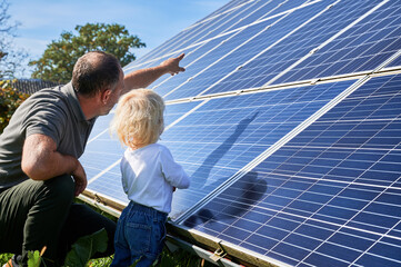 Man showing his small child the solar panels during sunny day. Father presenting to son modern energy resource. Back view of father and his kid watching on solar panels - obrazy, fototapety, plakaty