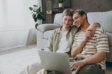 Young couple two gay IT men wear casual clothes work holding use laptop pc computer sit on sofa couch home hotel flat rest spend free spare time in living room Pride day june month love lgbtq concept