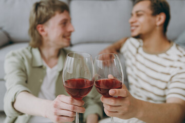 Young couple two gay men wear casual clothes together drink red wine sit on sofa couch at home flat rest spend free spare time in living room. Pride day june month love lgbtq concept. Focus on gasses.