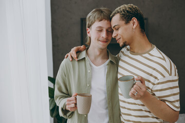 Young happy couple two gay men wear casual clothes together drink coffee close eyes hugging near window at home flat rest spend free spare time in living room Pride day june month love lgbtq concept.