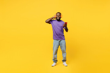 Fototapeta na wymiar Full body young man of African American ethnicity wear casual clothes purple t-shirt headphones listen music hold use mobile cell phone isolated on plain yellow background studio. Lifestyle concept.