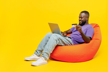 Full body young man of African American ethnicity in casual clothes purple t-shirt sit in bag chair using laptop pc computer hold credit bank card shopping online isolated on plain yellow background