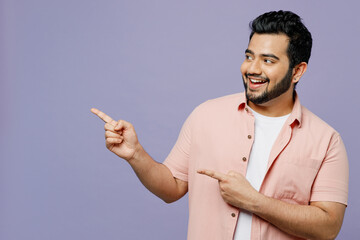 Young Indian man wear pink shirt white t-shirt casual clothes point index finger aside indicate on workspace area copy space mock up isolated on plain pastel light purple background studio portrait.