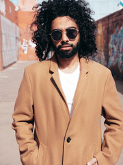 Handsome smiling hipster model.  Unshaven Arabian man dressed in brown coat clothes. Fashion male with long curly hairstyle posing outdoors in street. Cheerful and happy. In sunglasses. At sunny day