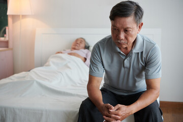 Fototapeta premium Depressed senior man deep in thoughts sitting on bed of his sick wife