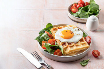 Savory breakfast with belgian waffles, fried egg and spinach, cherry tomato salad on pink tile background