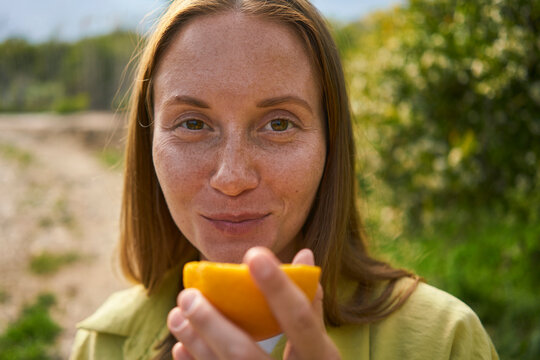 Smiling woman with freckles on face holding orange slice in orchard