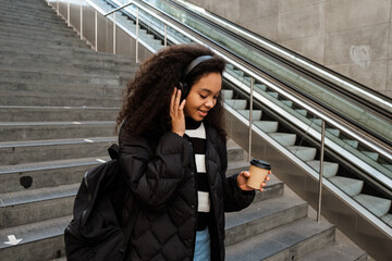 Obraz premium African girl listening music with headphones and holding takeaway coffee while going downstairs