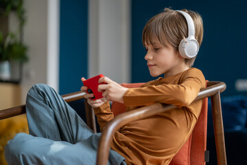 Children and screen time. Relaxed pre-teen boy child wearing headphones resting with smartphone at home, kid sitting on chair using mobile phone to listen to music or watch movies