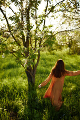 a beautiful, joyful woman is standing in a long orange dress, in the countryside, near a tree blooming with white flowers, during sunset, illuminated from the back and joyfully spinning