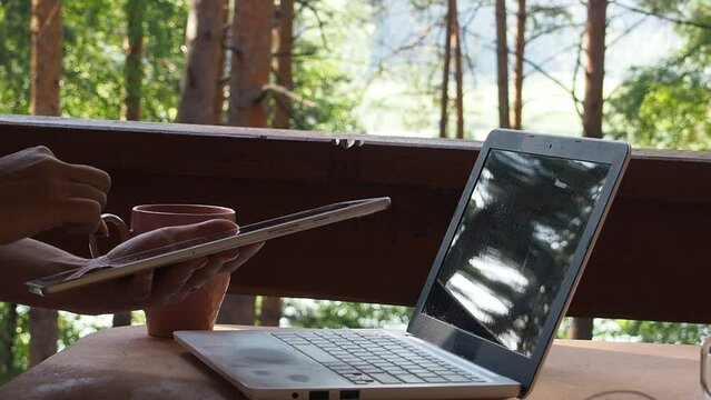 The girl uses modern technology on vacation in the forest. The girl works on a laptop, tablet and phone remotely. remote digital nomad on a table, mobile phone in nature with a green trees, freelance.